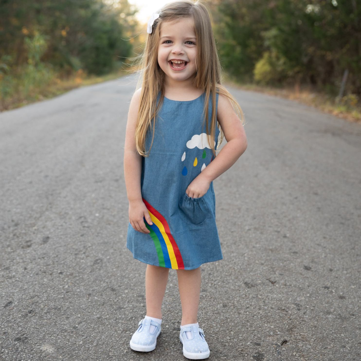 AL Limited Girls Blue Chambray Rainbow Overall Dress featuring a rainbow and cloud applique with yellow buttons on the shoulders.