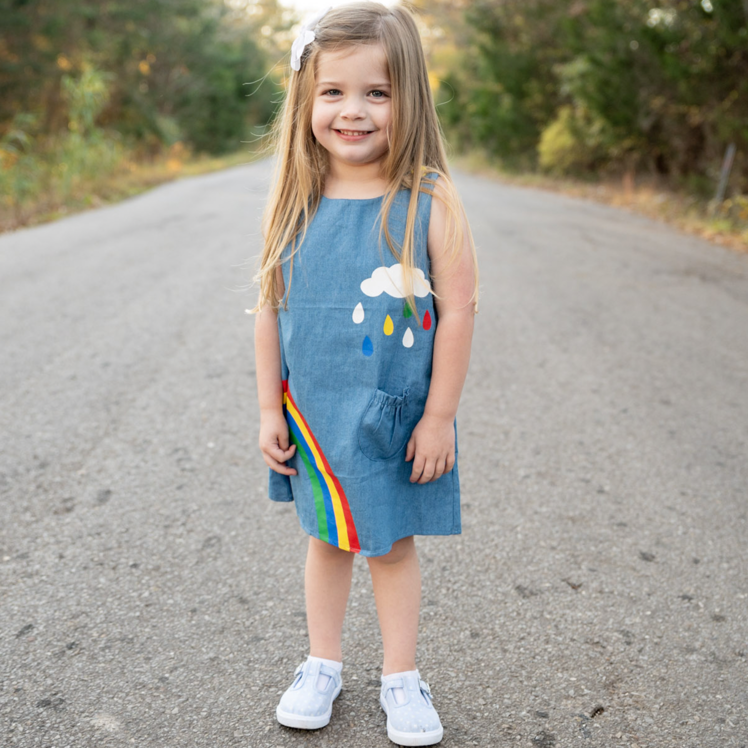 AL Limited Girls Blue Chambray Rainbow Overall Dress featuring a rainbow and cloud applique with yellow buttons on the shoulders.