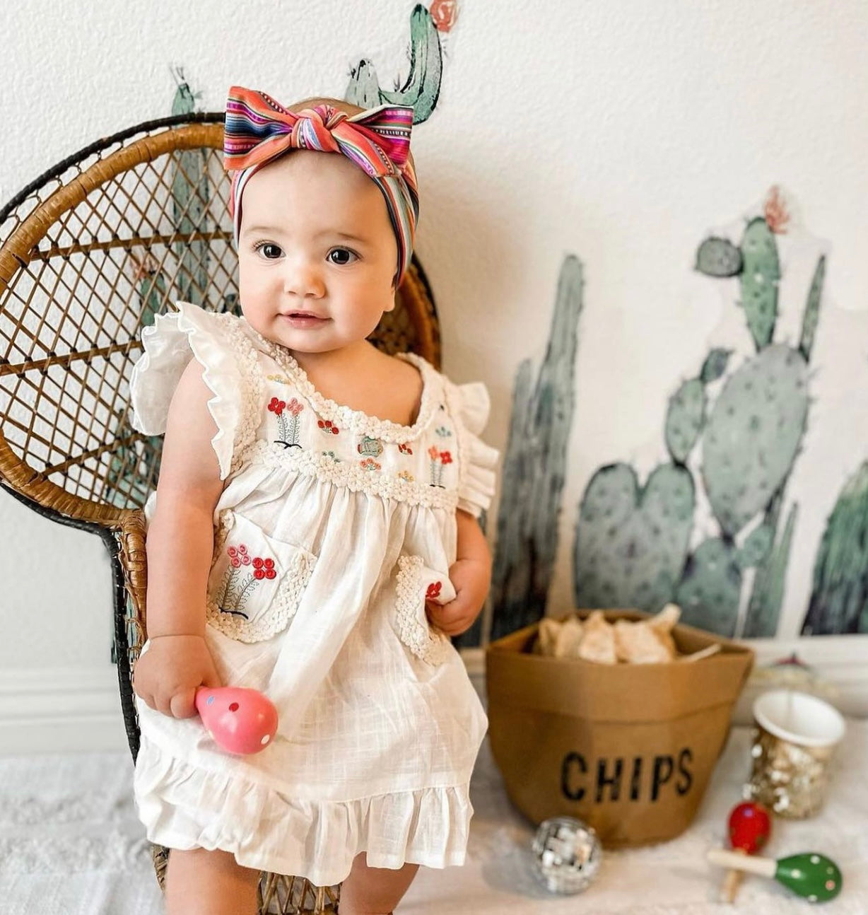 A white Maribel embroidered dress featuring colorful embroidery on the front pockets and chest, with ribbon detailing along the edges.