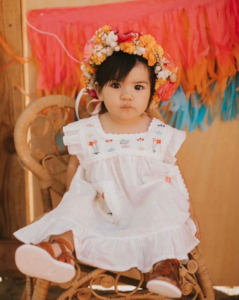 A white Maribel embroidered dress featuring colorful embroidery on the front pockets and chest, with ribbon detailing along the edges.