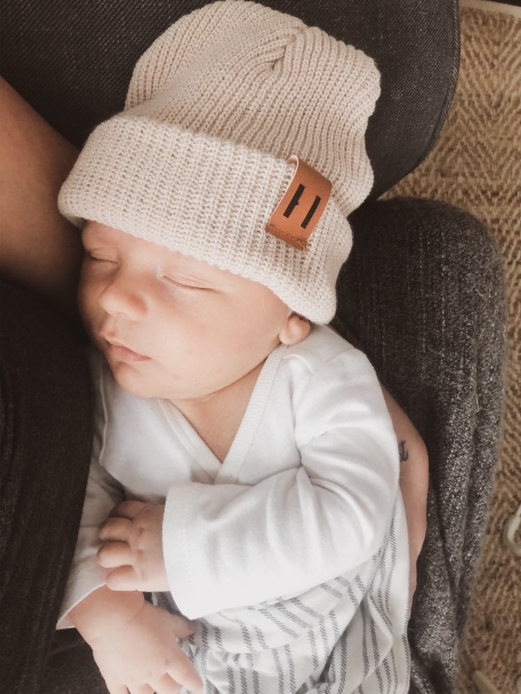 A soft and stylish neutral knit bonnet for babies, designed to fit ages 6M to 18M, featuring a secure brown tag.