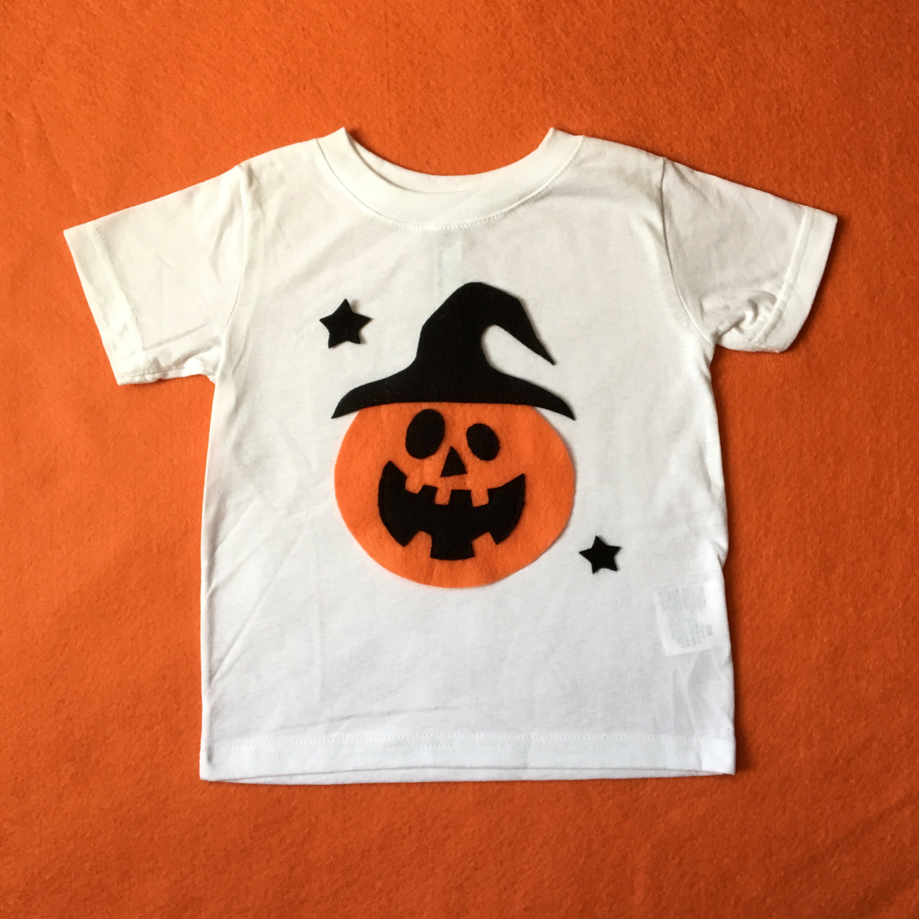 A handmade Pumpkin Witch Kids Tee featuring a cute pumpkin design, perfect for Halloween celebrations, made with soft fabric and appliquéd felt pieces.