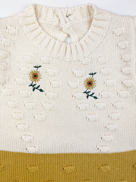 A cozy sunflower knitted onesie in gold and cream, featuring crochet sunflower details and wooden buttons for easy diaper changes.