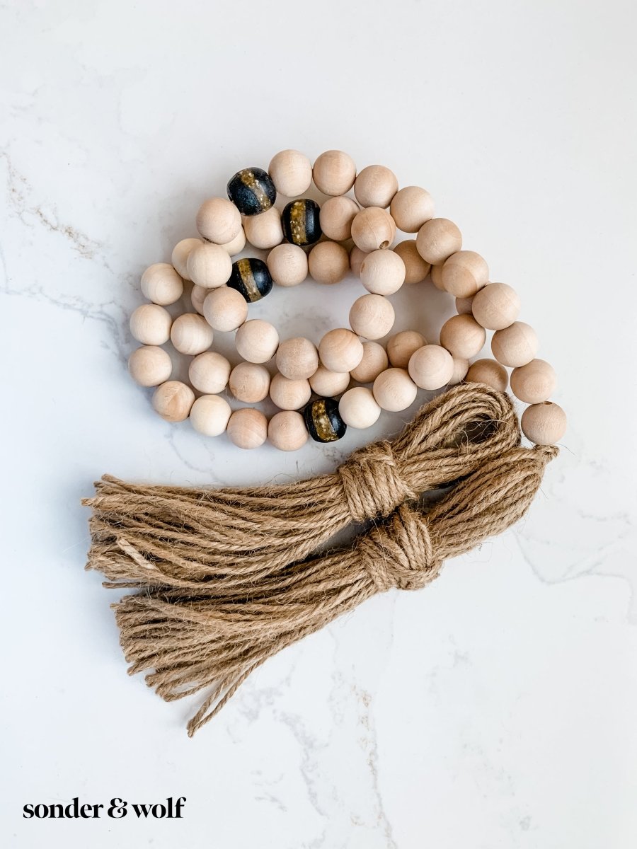 A 4ft handmade wood bead garland featuring light unfinished wood beads and four distinctive Black Kente Krobo beads, adorned with jute tassels.