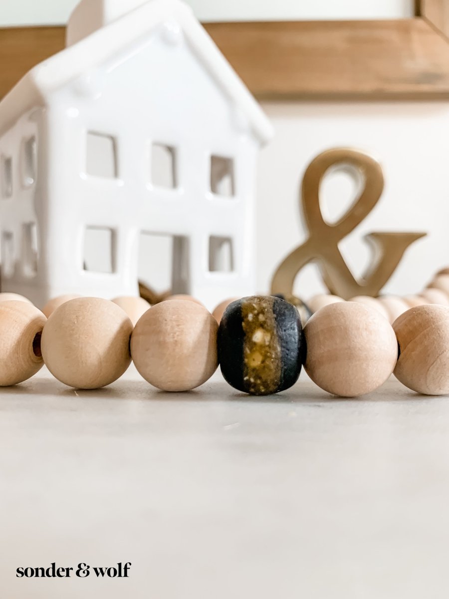 A 4ft handmade wood bead garland featuring light unfinished wood beads and four distinctive Black Kente Krobo beads, adorned with jute tassels.