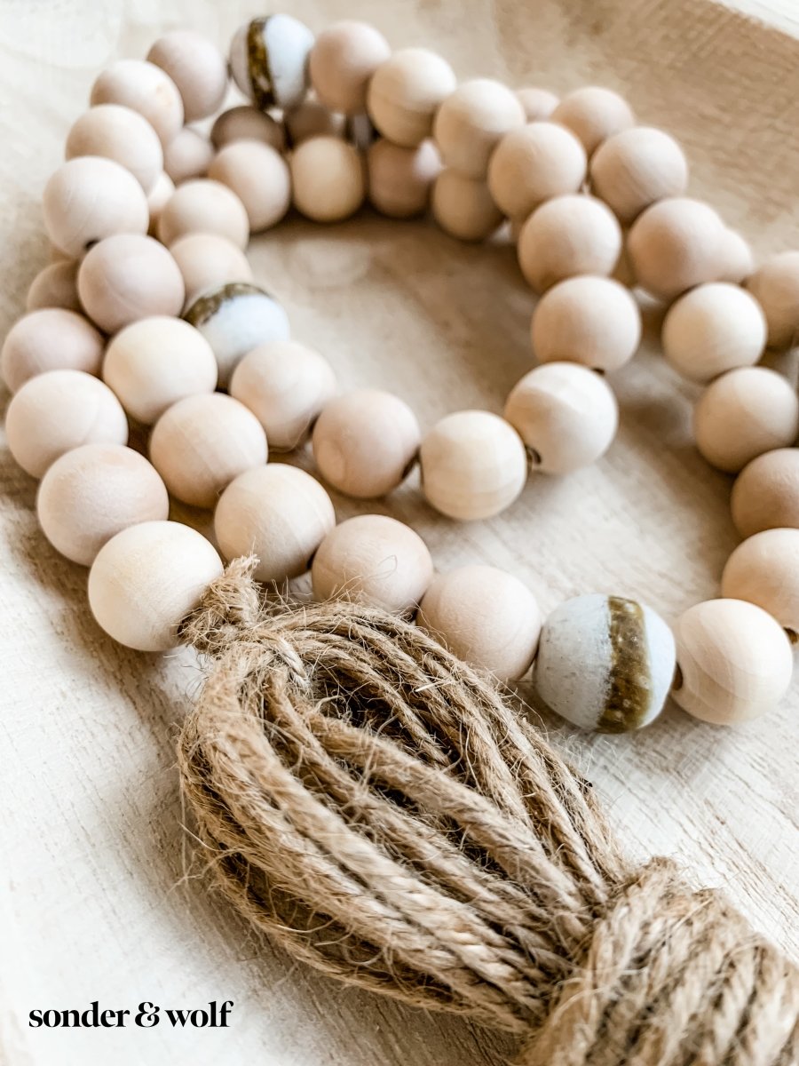 A 4ft handmade wood bead garland featuring light unfinished wood beads and four distinctive White Kente Krobo beads, adorned with jute tassels.