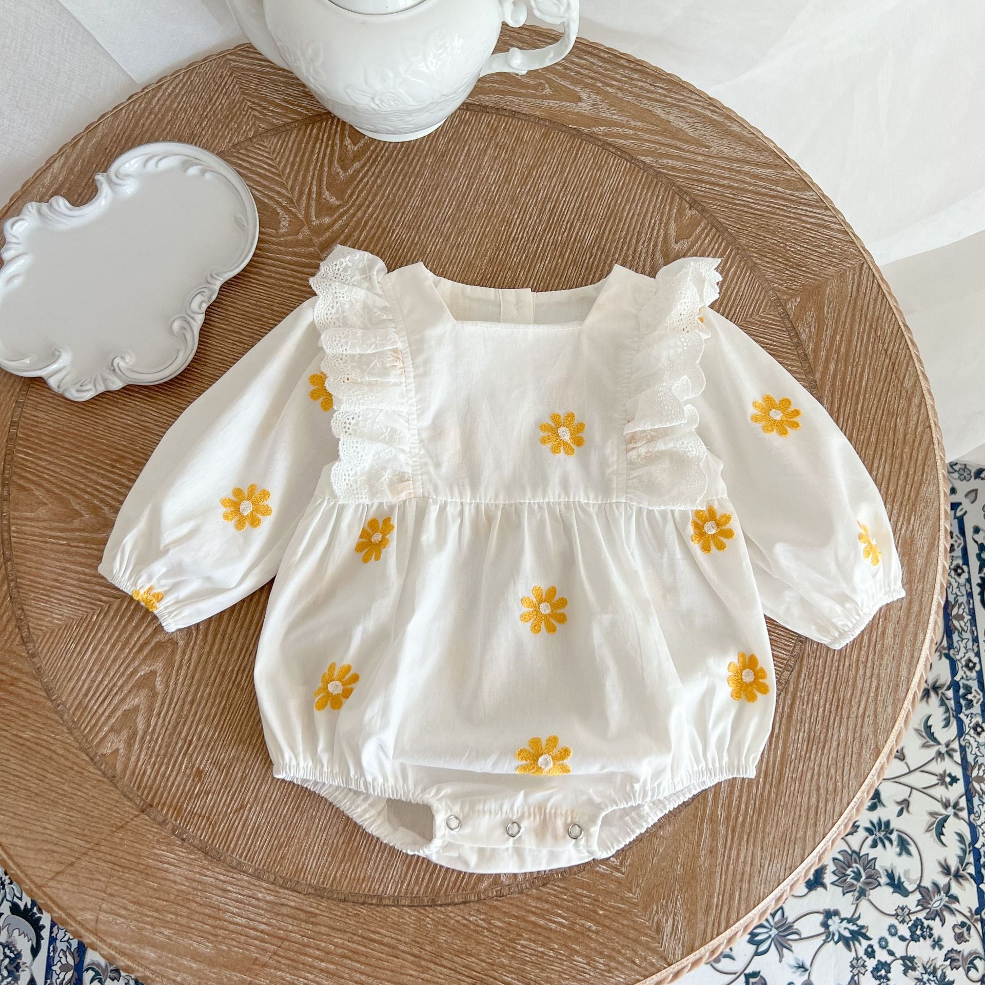 A baby girl wearing a floral embroidered onesie with ruffle design and square collar, showcasing a stylish and comfortable look.