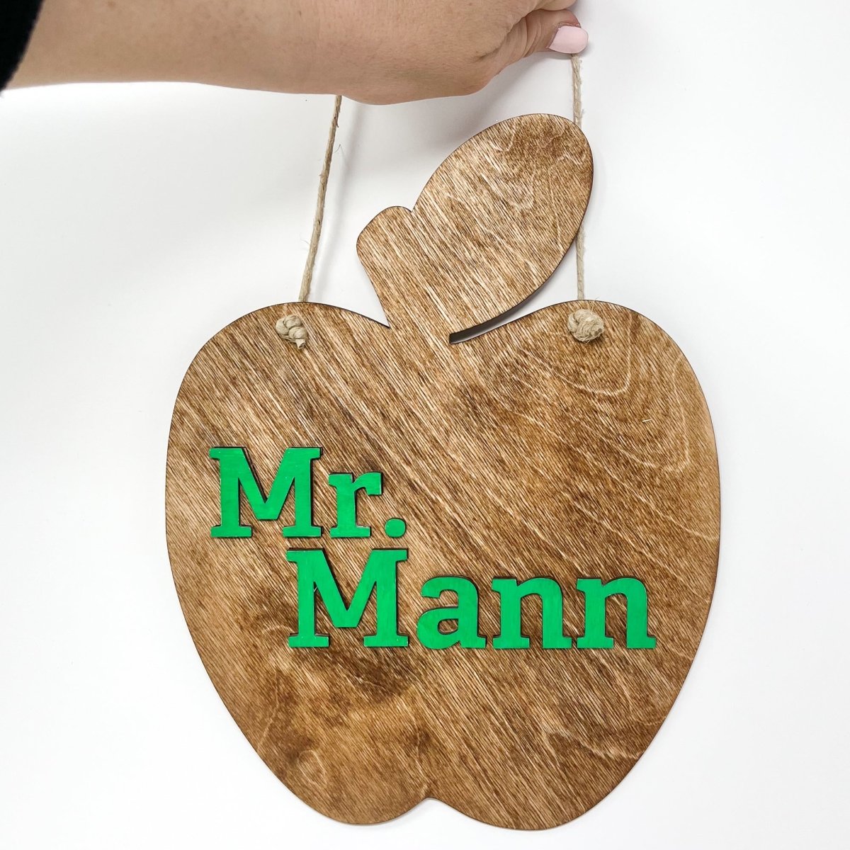 A personalized teacher apple sign made of natural wood, featuring raised lettering and colorful design, ready to hang with jute rope.