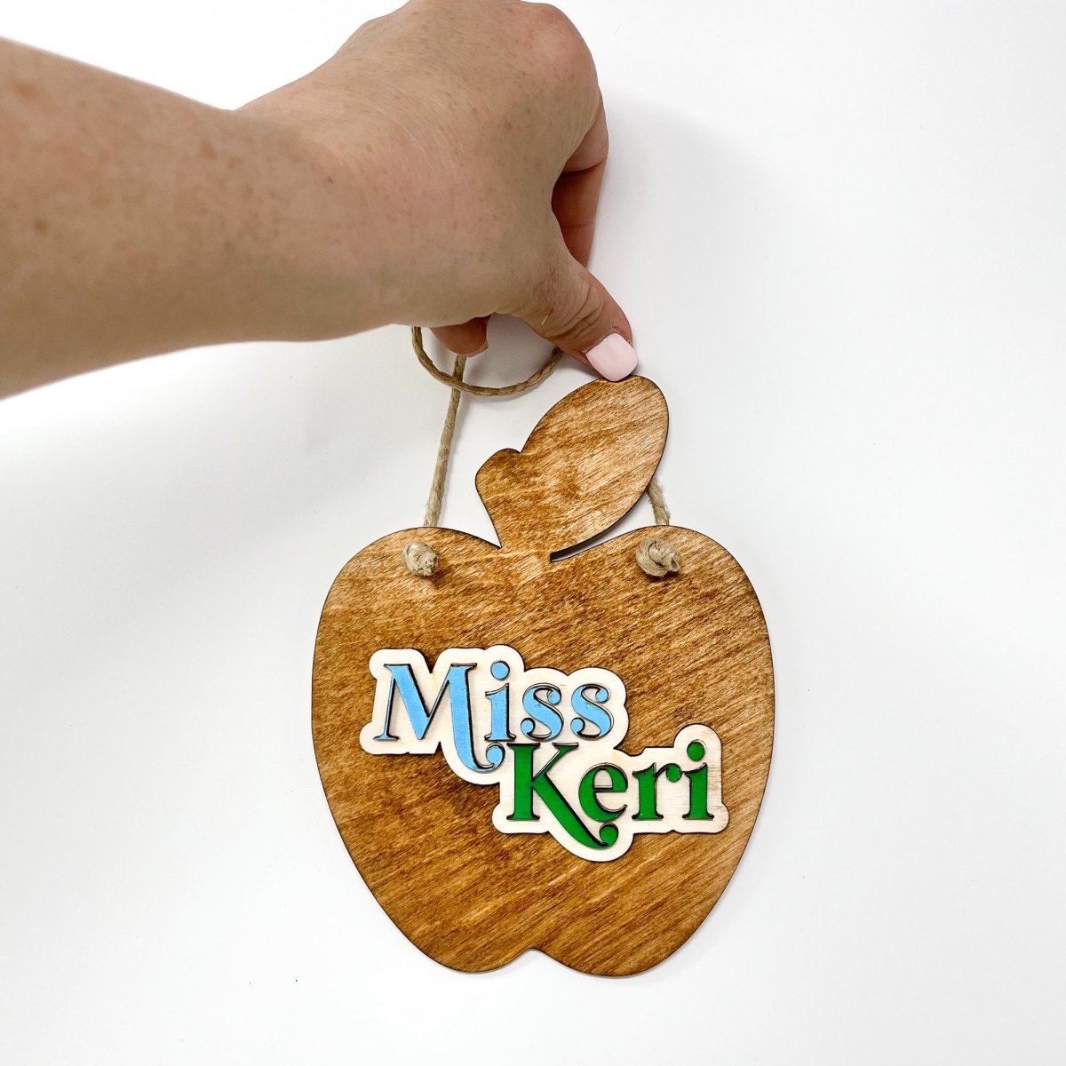 A personalized teacher apple sign made of natural wood, featuring raised lettering and colorful design, ready to hang with jute rope.