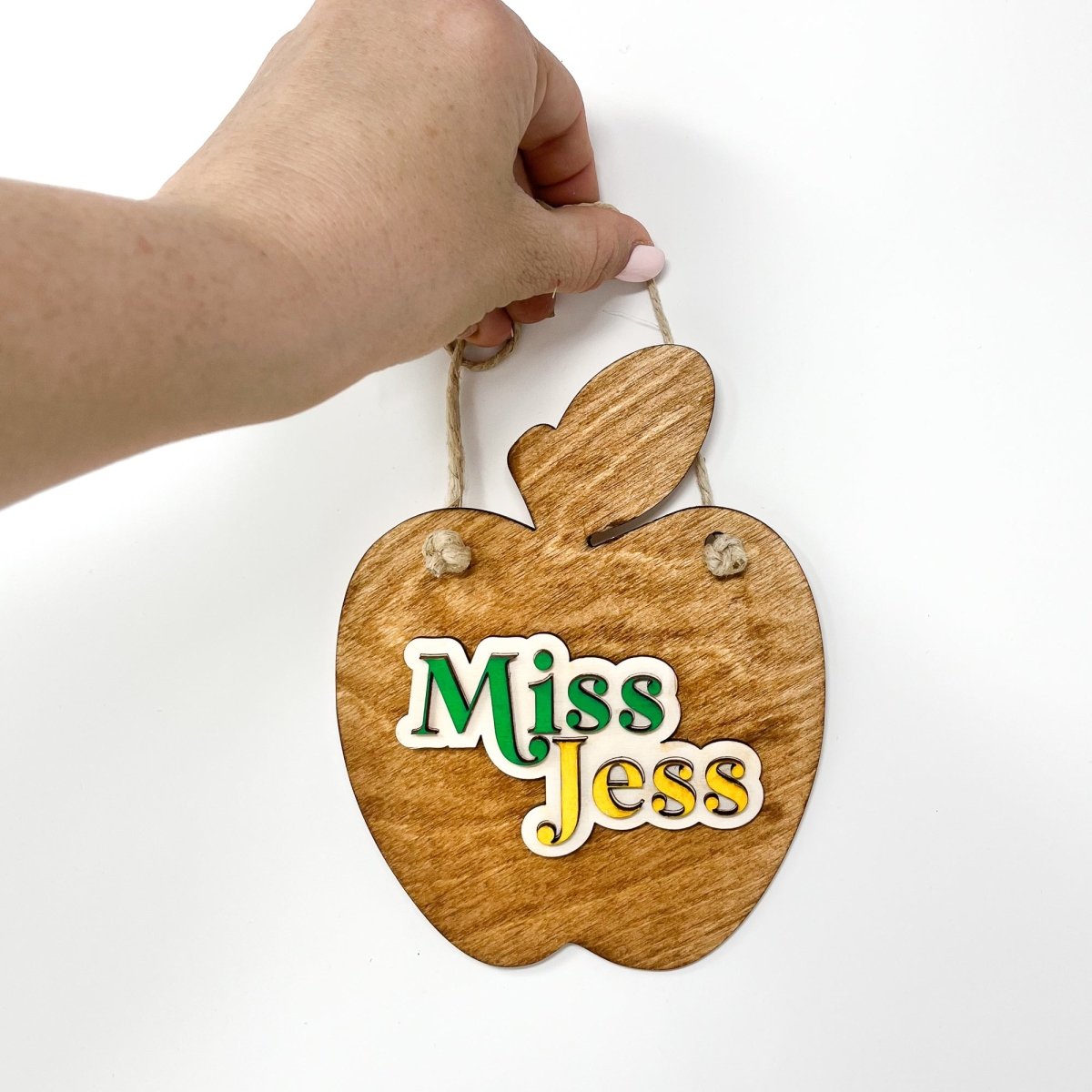 A personalized teacher apple sign made of natural wood, featuring raised lettering and colorful design, ready to hang with jute rope.