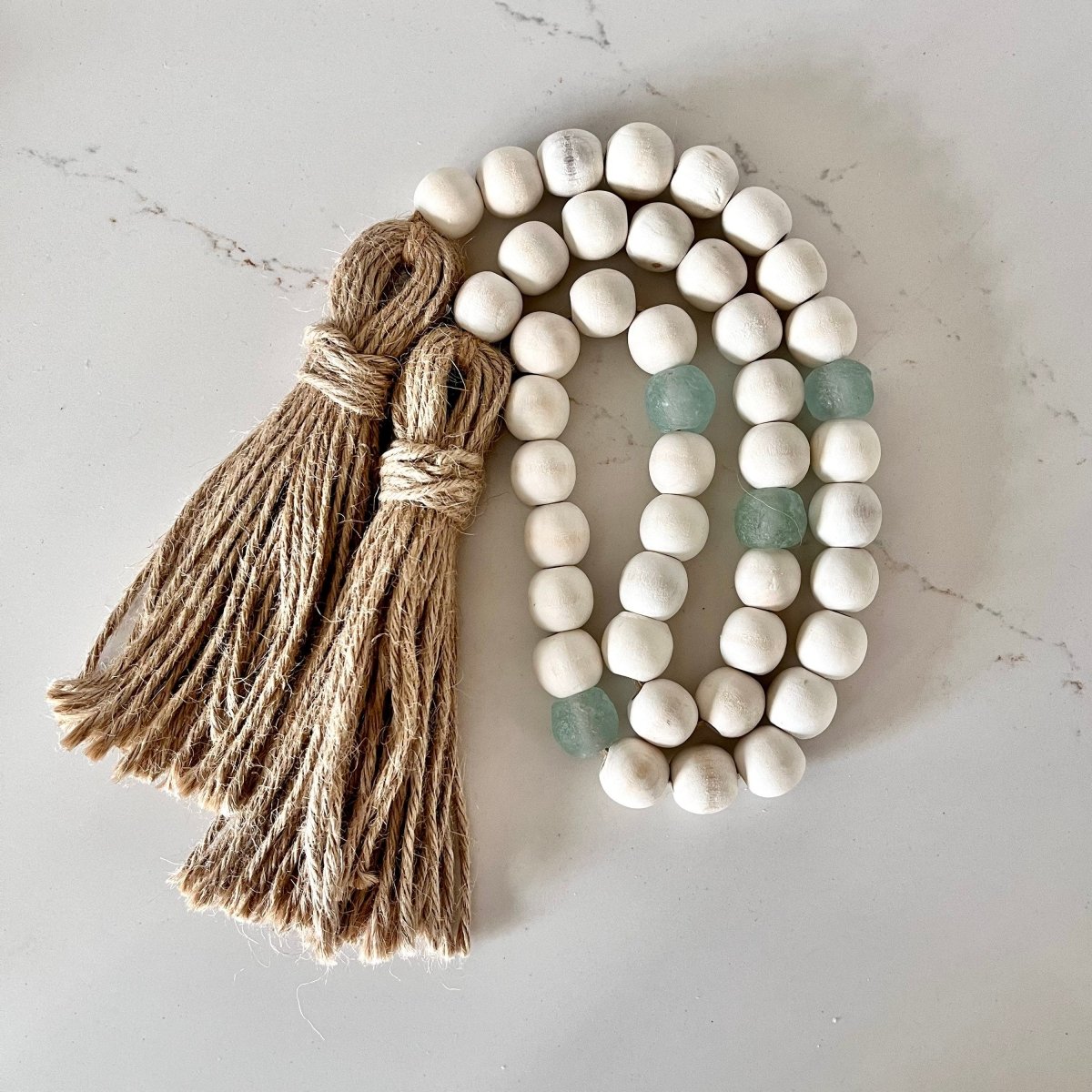 A beautiful whitewashed wood bead garland featuring aqua recycled glass beads and jute tassels, perfect for coastal decor.