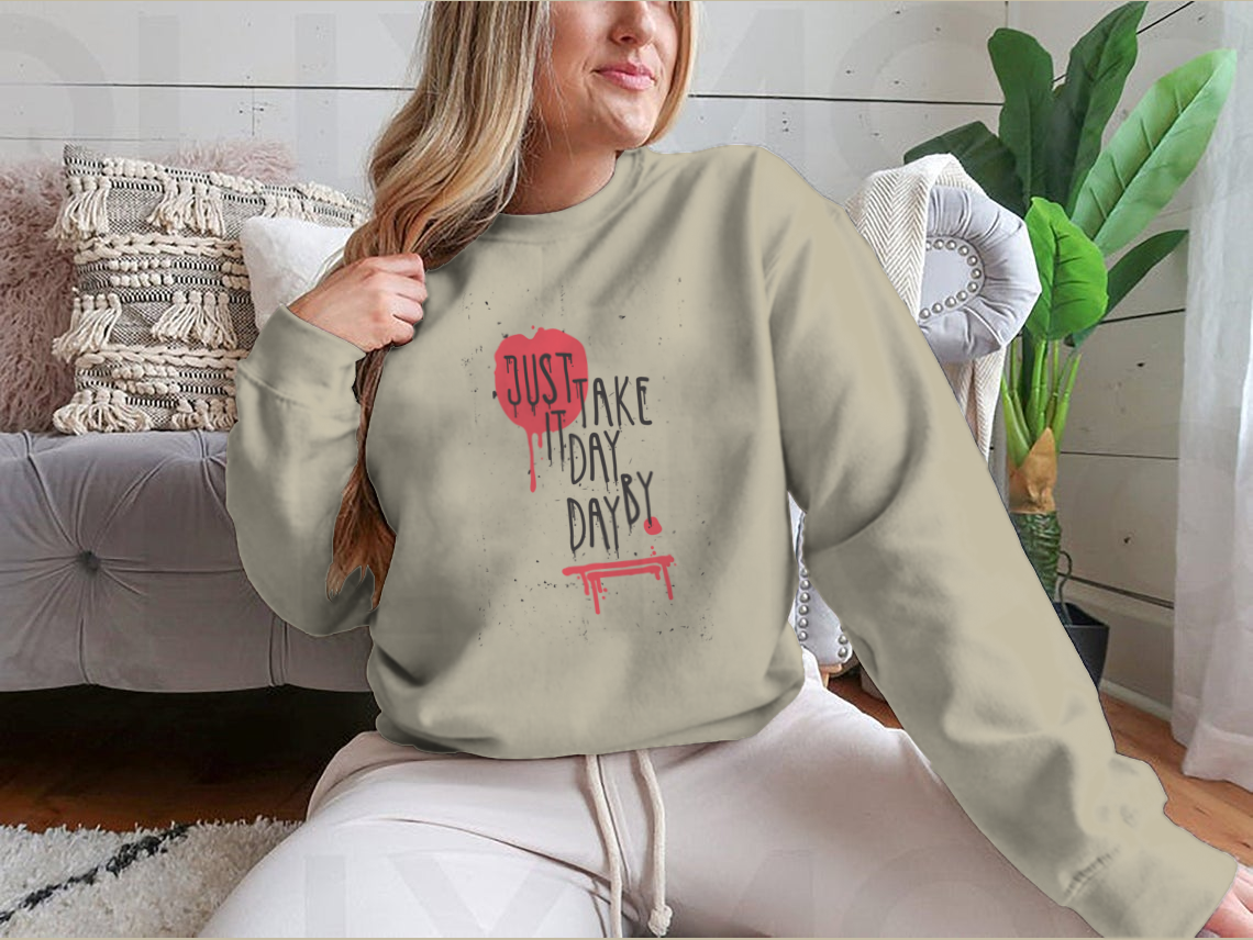 A cozy sweatshirt featuring the motivational phrase 'Just Take It Day By Day' in stylish typography, perfect for casual wear.