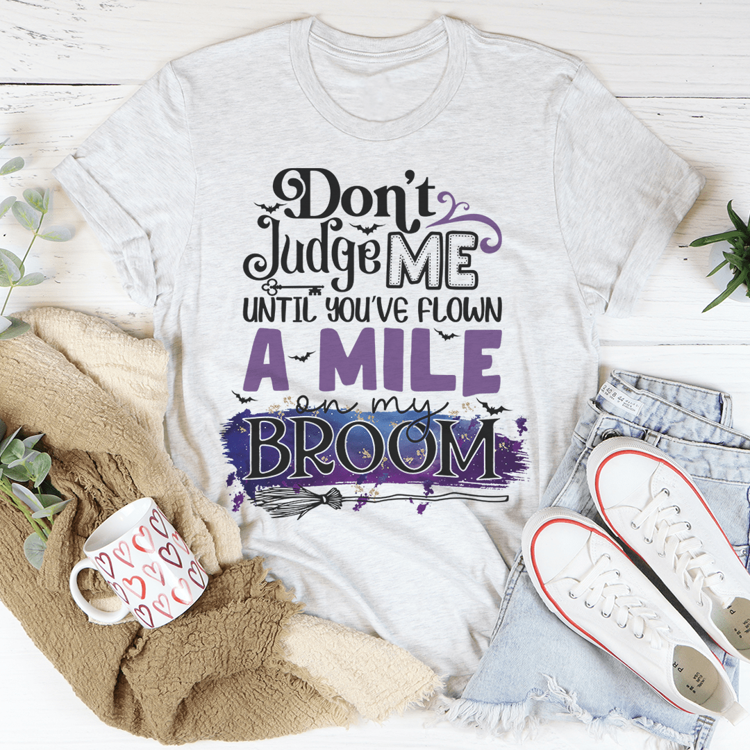 A stylish black t-shirt featuring the phrase 'Don't Judge Me Until You've Flown A Mile On My Broom' printed in a whimsical font.