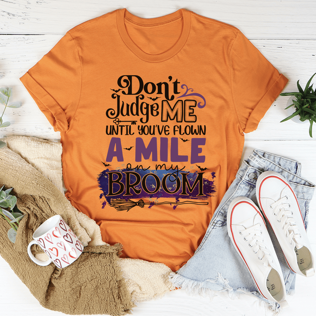 A stylish black t-shirt featuring the phrase 'Don't Judge Me Until You've Flown A Mile On My Broom' printed in a whimsical font.