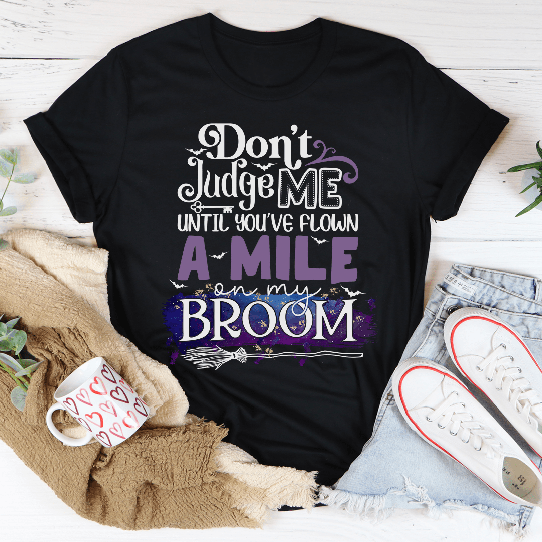 A stylish black t-shirt featuring the phrase 'Don't Judge Me Until You've Flown A Mile On My Broom' printed in a whimsical font.