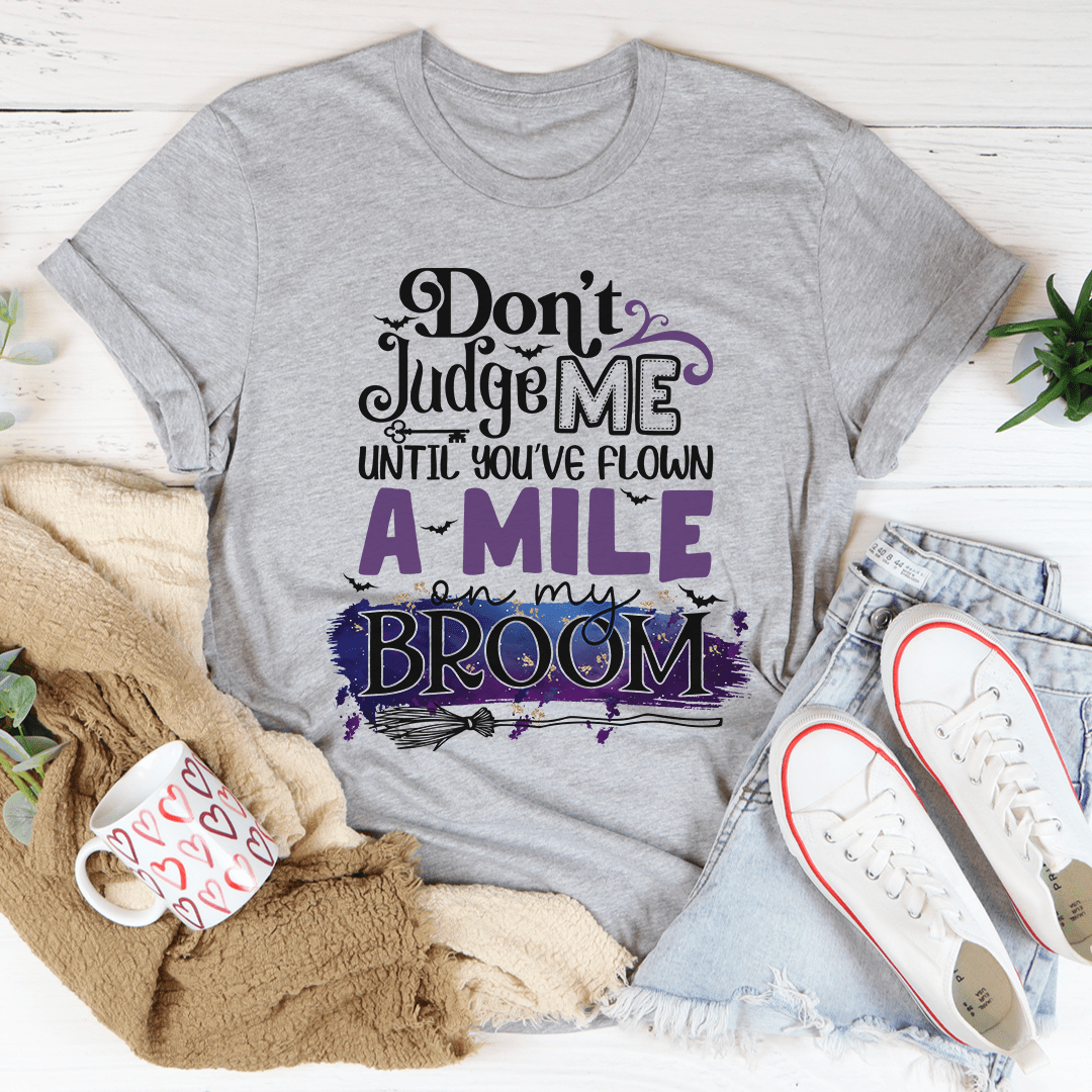 A stylish black t-shirt featuring the phrase 'Don't Judge Me Until You've Flown A Mile On My Broom' printed in a whimsical font.