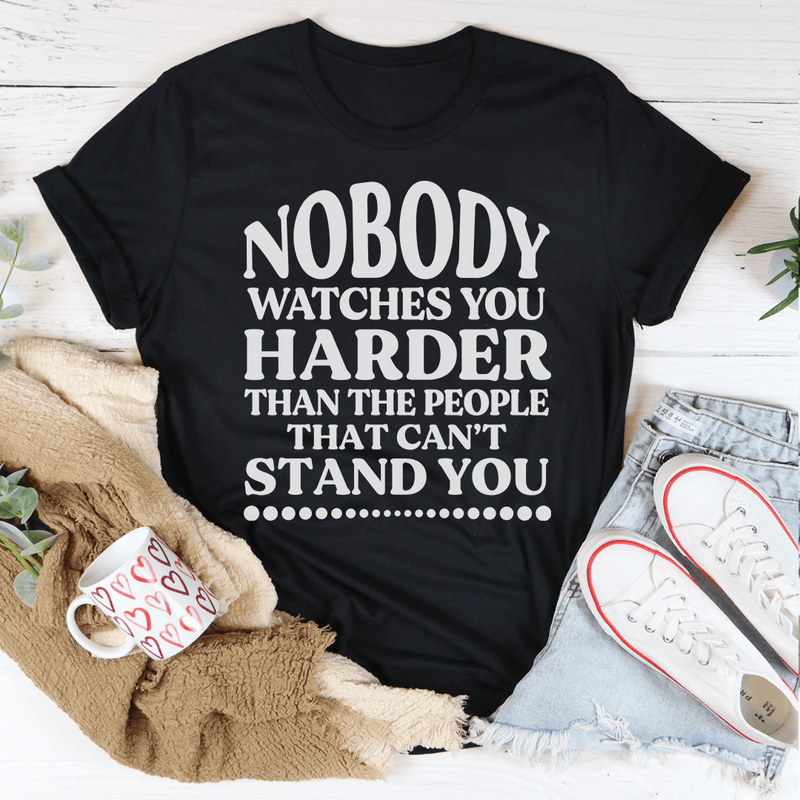 A stylish black t-shirt featuring the phrase 'Nobody Watches You Harder Than The People That Can't Stand You' in bold white lettering.