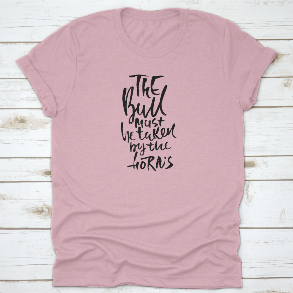 A stylish t-shirt featuring hand-drawn dry brush lettering with the phrase 'The Bull Must Be Taken By The Horns', displayed on a model.