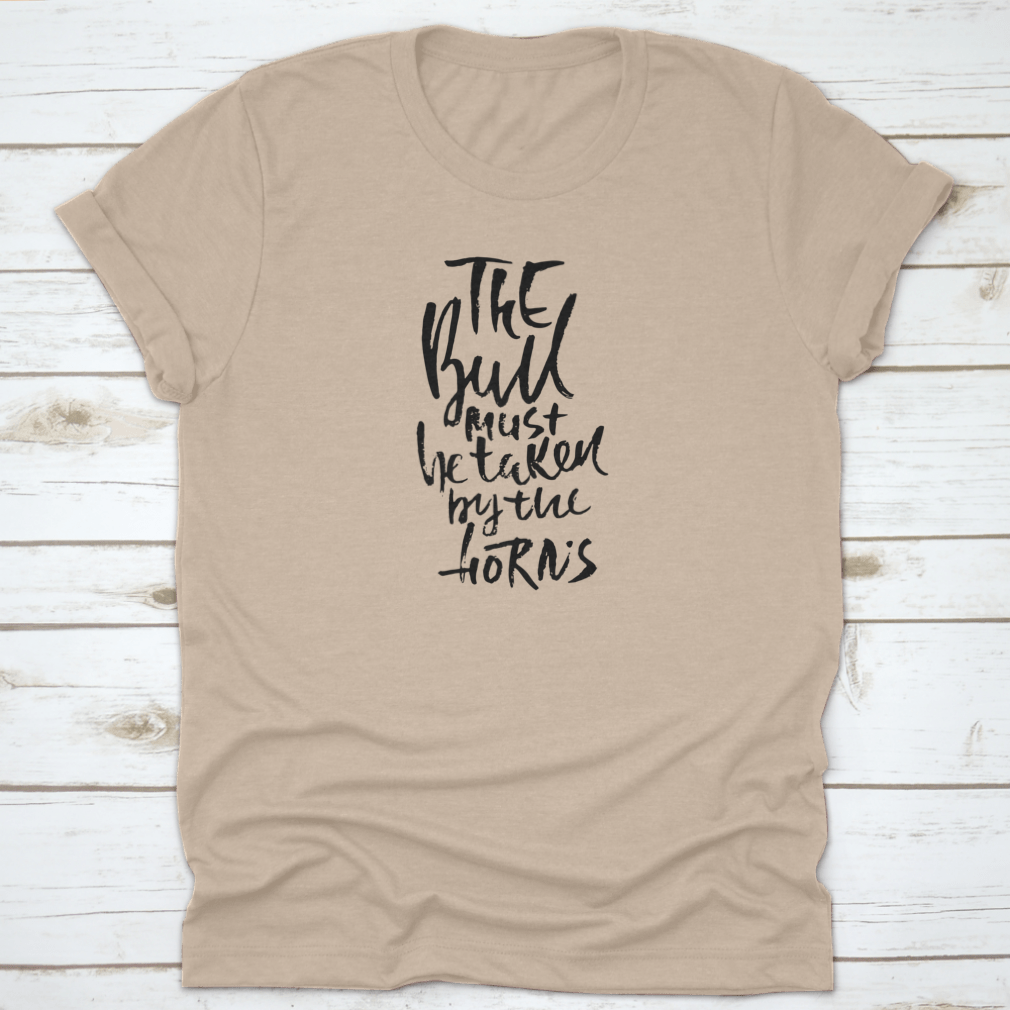 A stylish t-shirt featuring hand-drawn dry brush lettering with the phrase 'The Bull Must Be Taken By The Horns', displayed on a model.