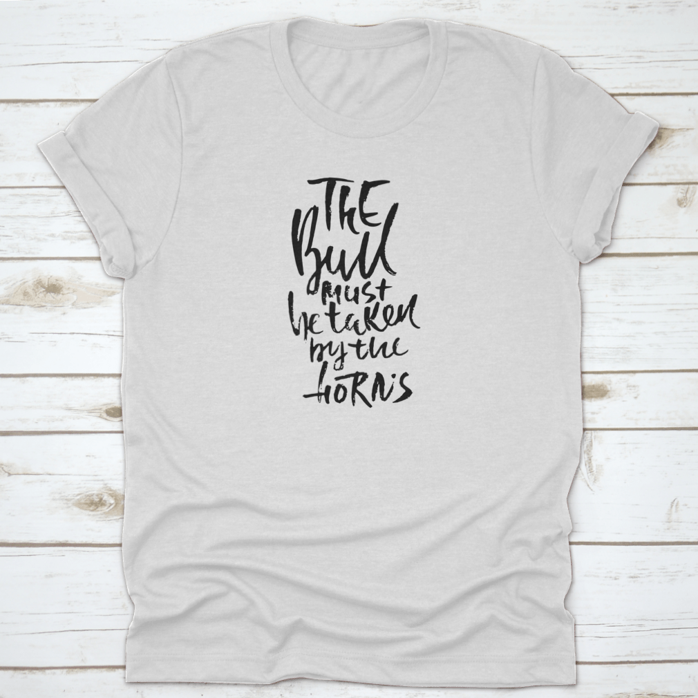 A stylish t-shirt featuring hand-drawn dry brush lettering with the phrase 'The Bull Must Be Taken By The Horns', displayed on a model.