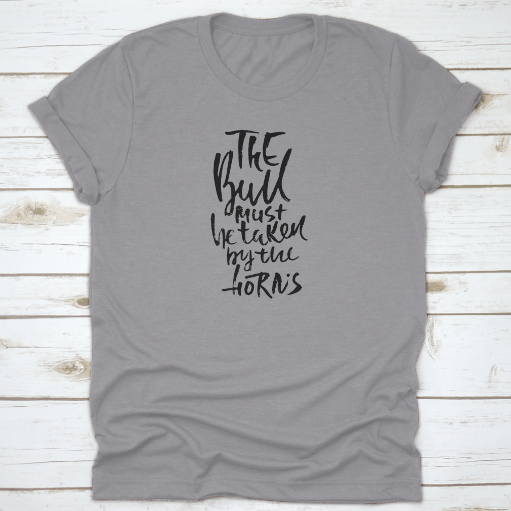 A stylish t-shirt featuring hand-drawn dry brush lettering with the phrase 'The Bull Must Be Taken By The Horns', displayed on a model.