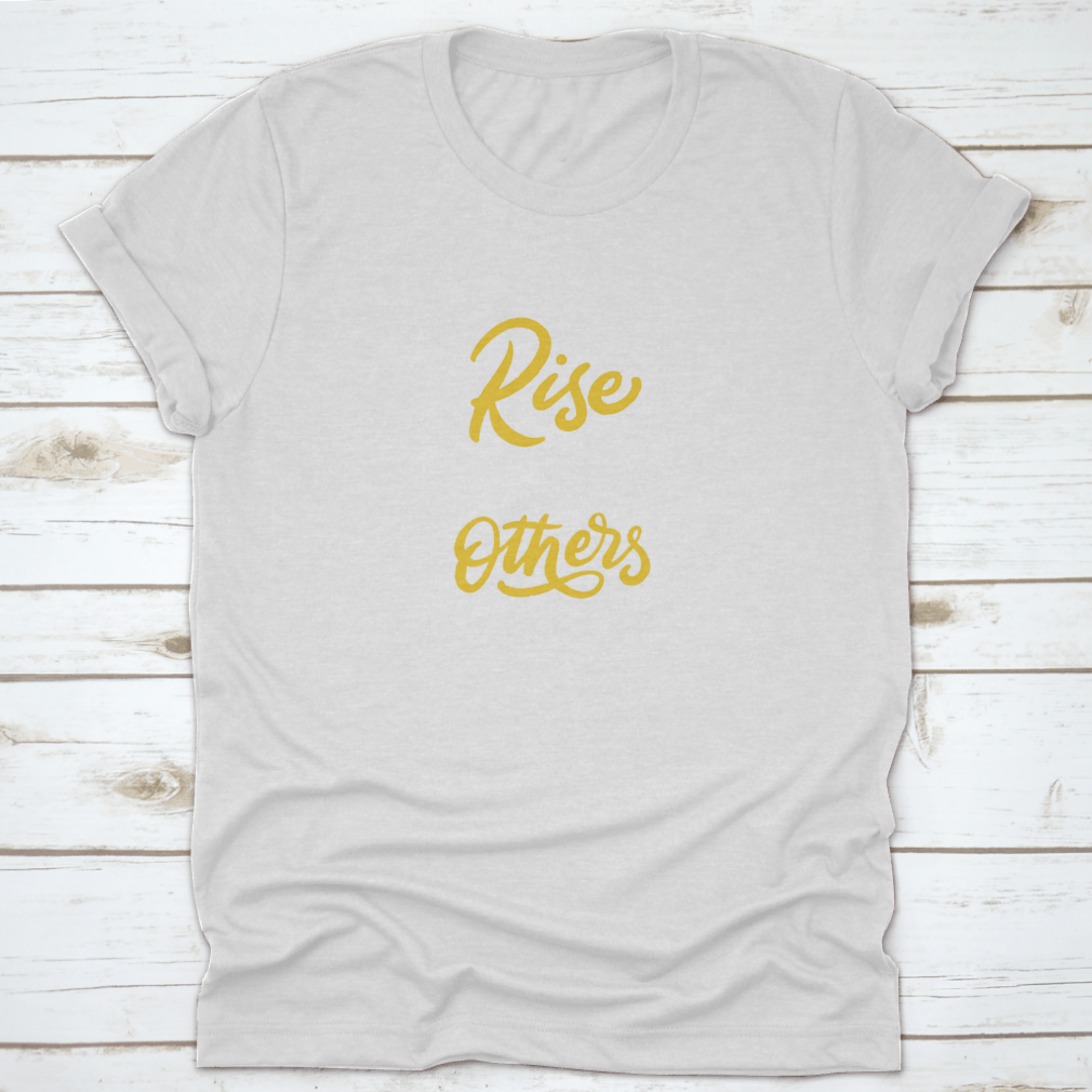 A motivational T-shirt featuring the quote 'We Rise By Lifting Others', made from soft cotton fabric, displayed on a neutral background.