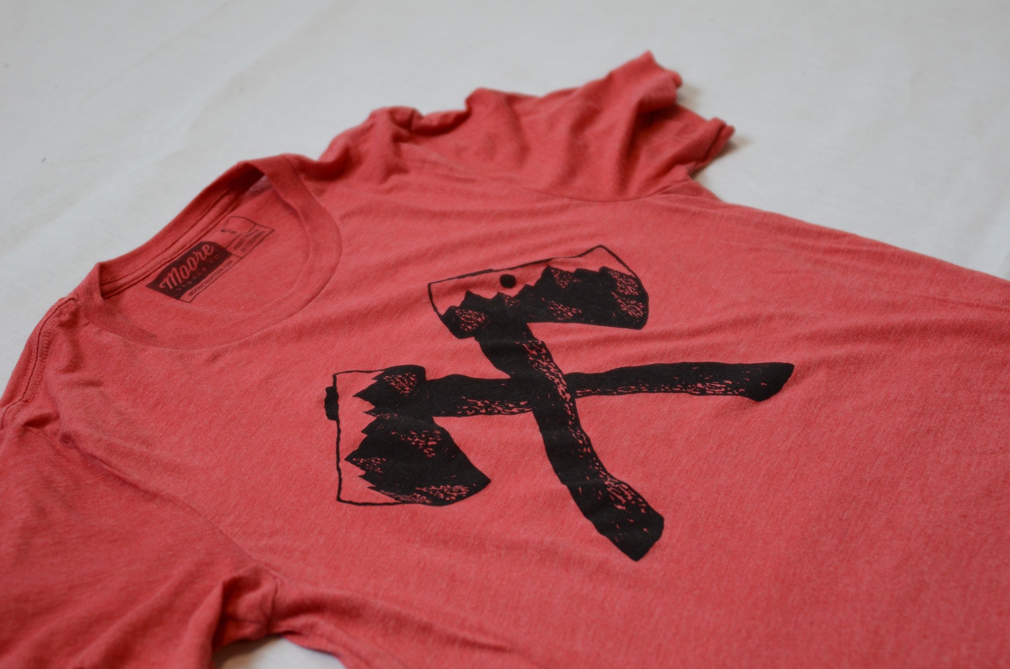 Axe Tee in red featuring white water-based ink screen print, made from 100% ringspun cotton, displayed on a neutral background.