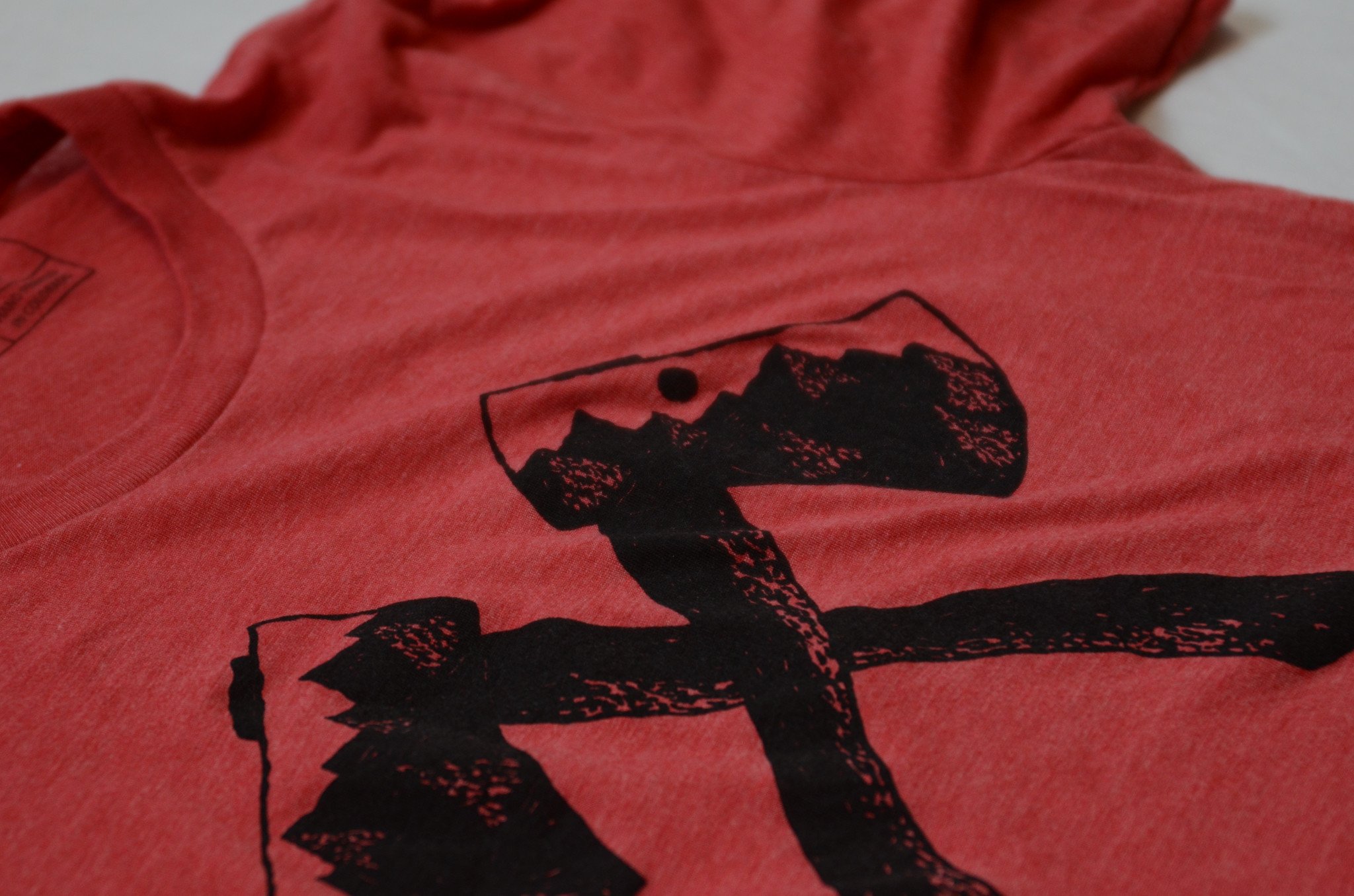 Axe Tee in red featuring white water-based ink screen print, made from 100% ringspun cotton, displayed on a neutral background.