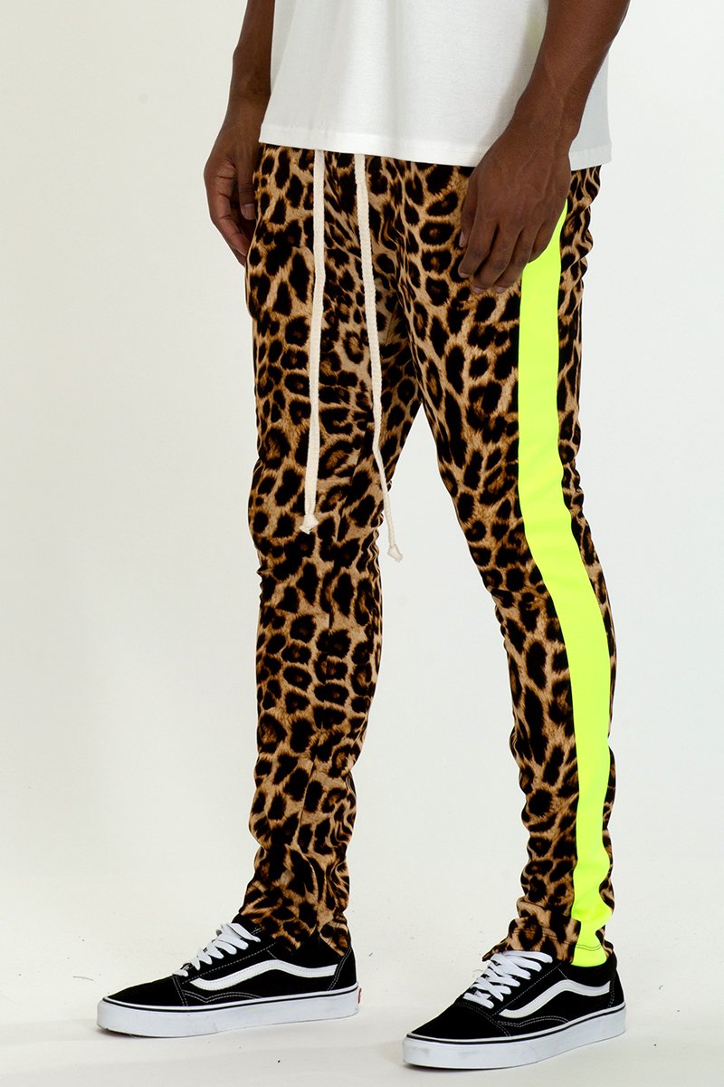 Cheetah print track pants with lime stripe detail, featuring an elastic waist and hidden ankle zippers, displayed on a model.