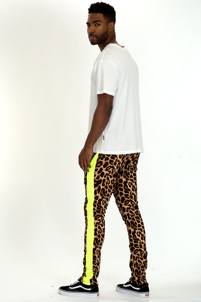 Cheetah print track pants with lime stripe detail, featuring an elastic waist and hidden ankle zippers, displayed on a model.