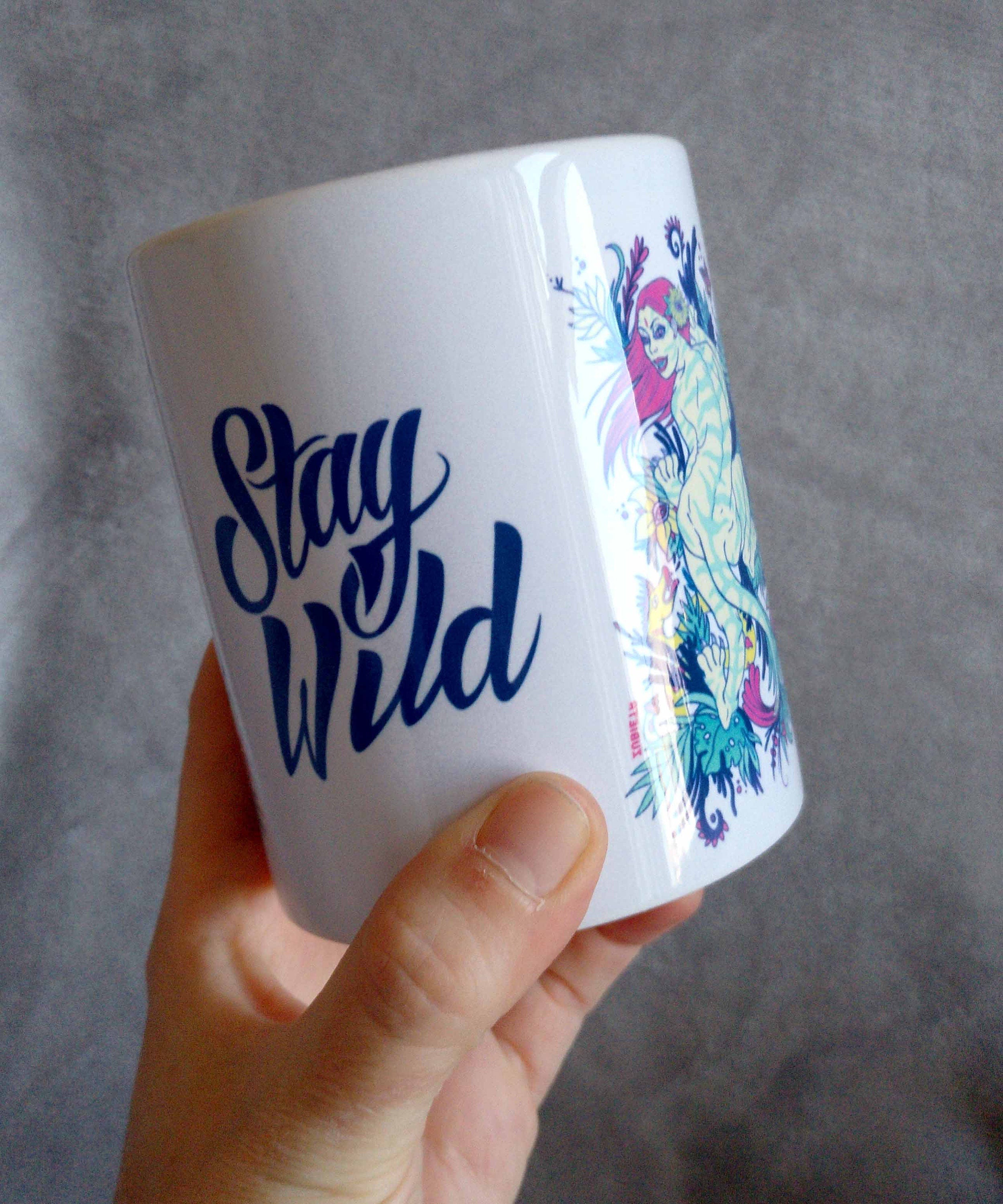 Ceramic mug featuring a vibrant tiger woman design with 'Stay Wild' lettering in the center, showcasing artistic flair.