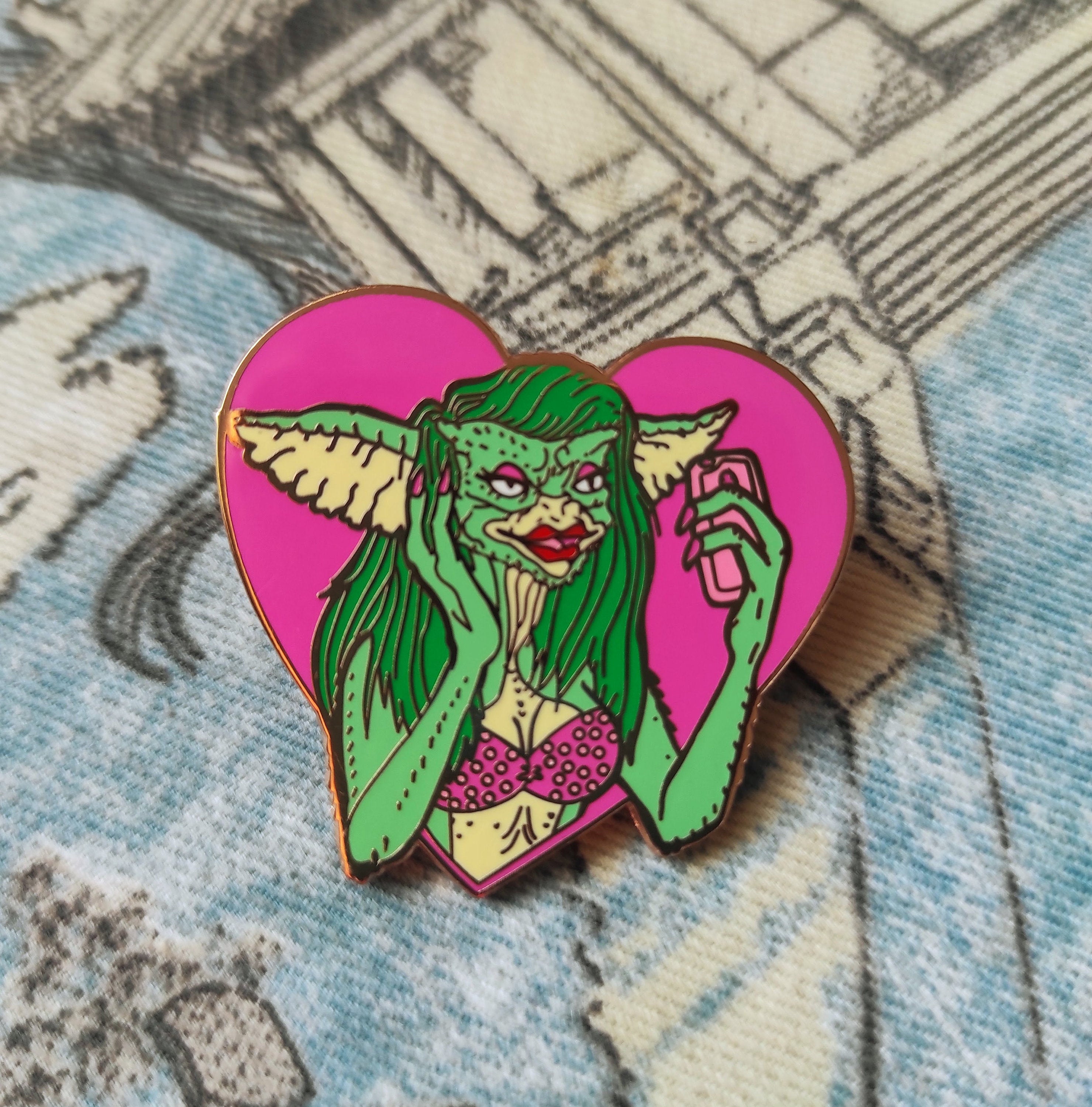 Greta the Gremlin Maxi Pin featuring a cute and creepy design, 2 inches wide, made of hard enamel with a rose gold metal finish.