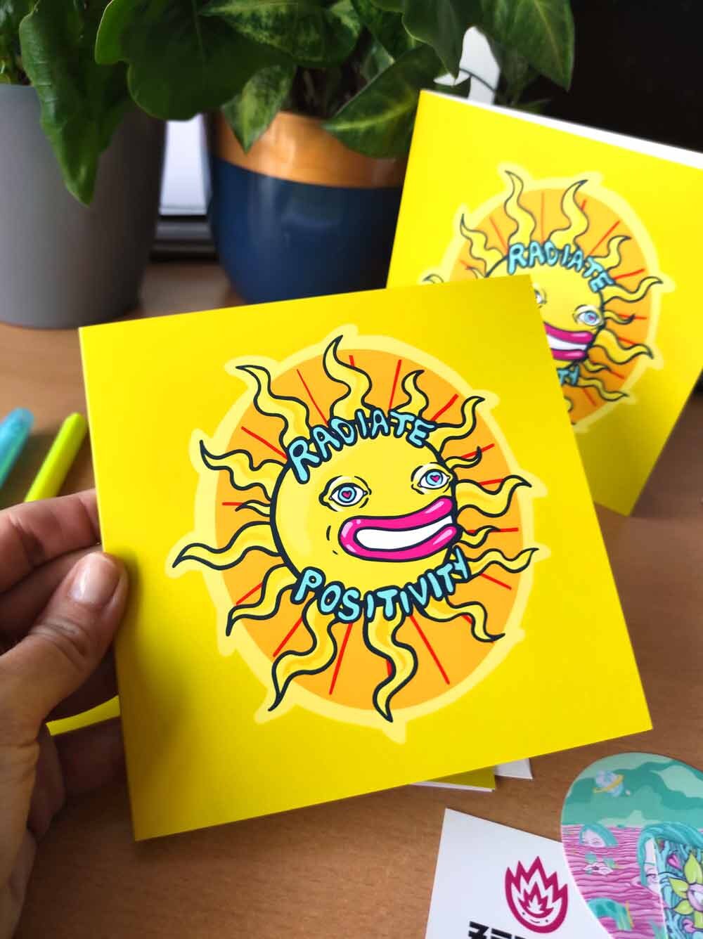 A vibrant psychedelic card titled 'Radiate Positivity', featuring colorful designs symbolizing love and friendship, with a square envelope and sticker.