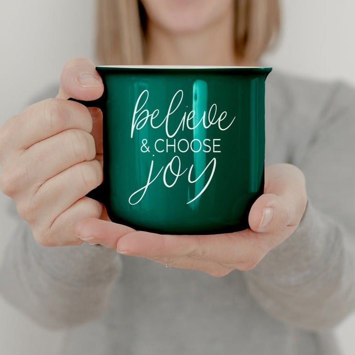 A ceramic Believe & Joy Mug in red with white lettering, featuring a positive message on both sides, perfect for holiday gifting.