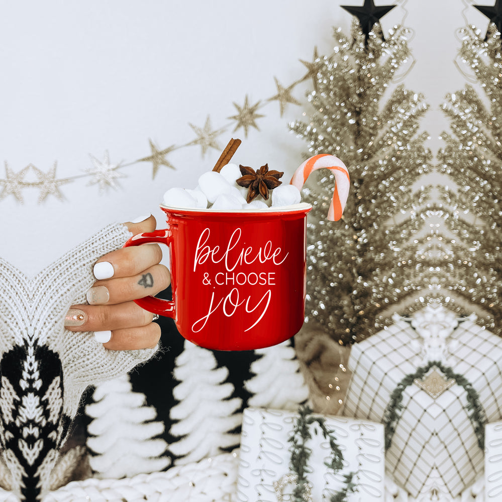 A ceramic Believe & Joy Mug in red with white lettering, featuring a positive message on both sides, perfect for holiday gifting.