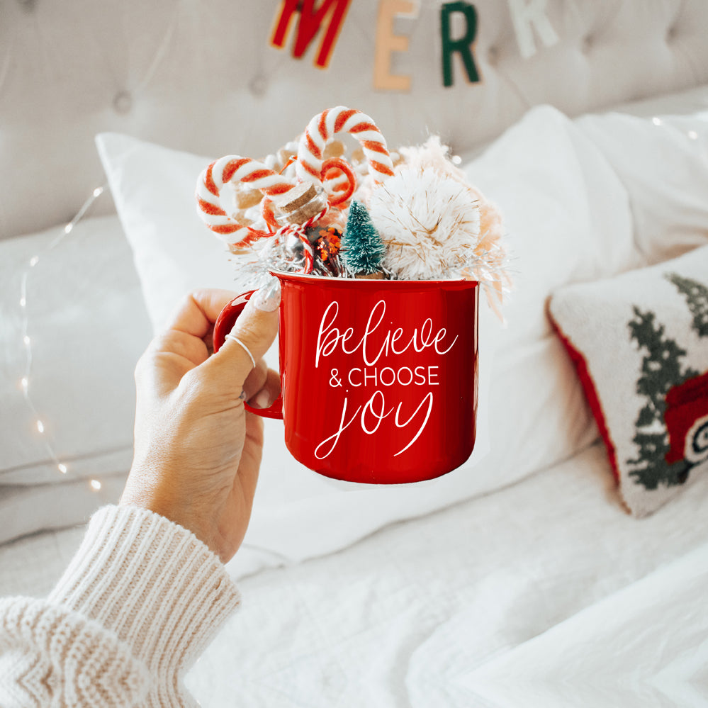 A ceramic Believe & Joy Mug in red with white lettering, featuring a positive message on both sides, perfect for holiday gifting.