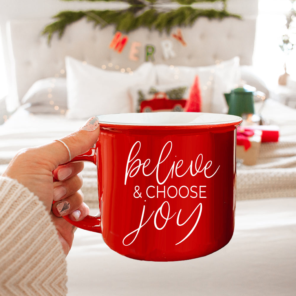 A ceramic Believe & Joy Mug in red with white lettering, featuring a positive message on both sides, perfect for holiday gifting.