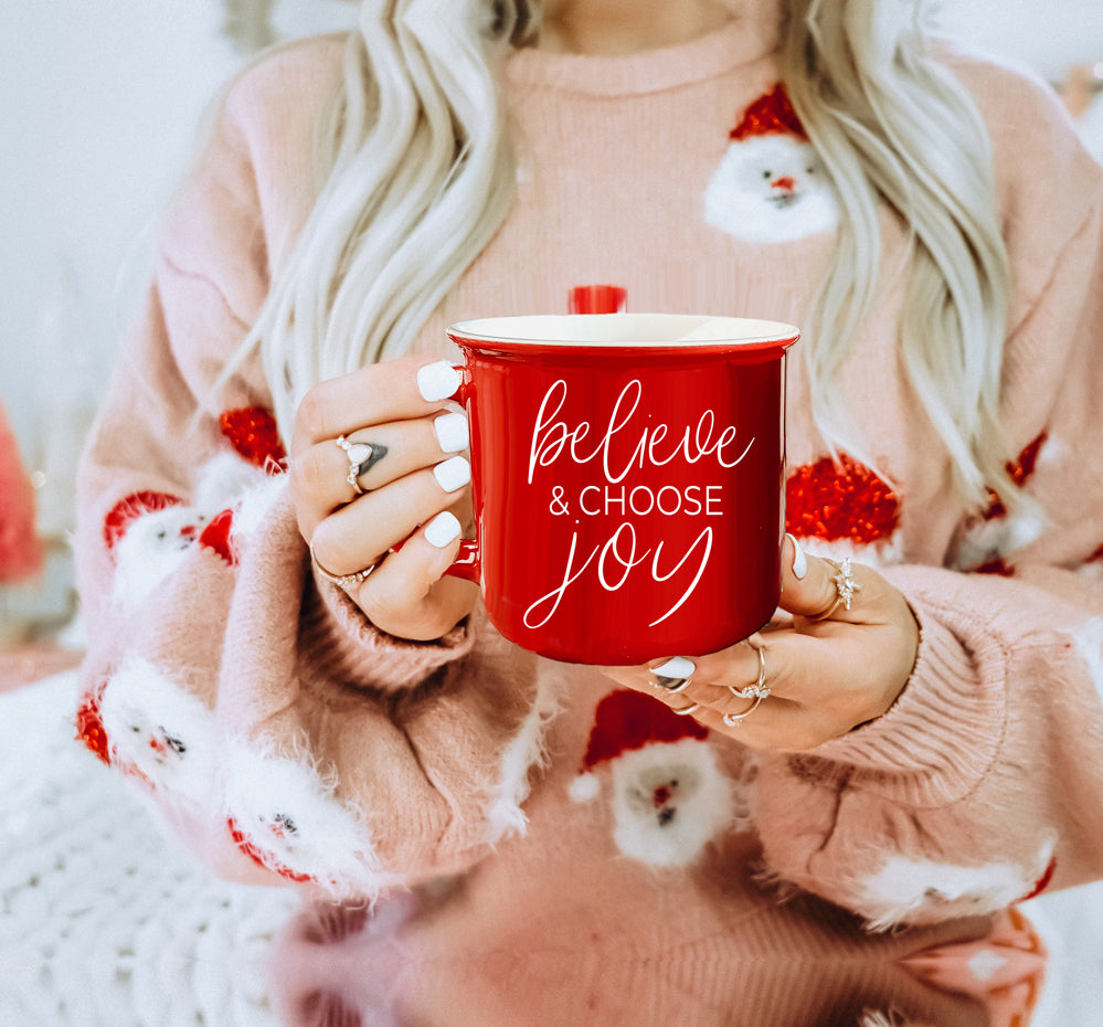 A ceramic Believe & Joy Mug in red with white lettering, featuring a positive message on both sides, perfect for holiday gifting.