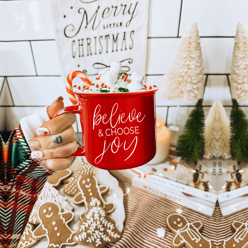 A ceramic Believe & Joy Mug in red with white lettering, featuring a positive message on both sides, perfect for holiday gifting.