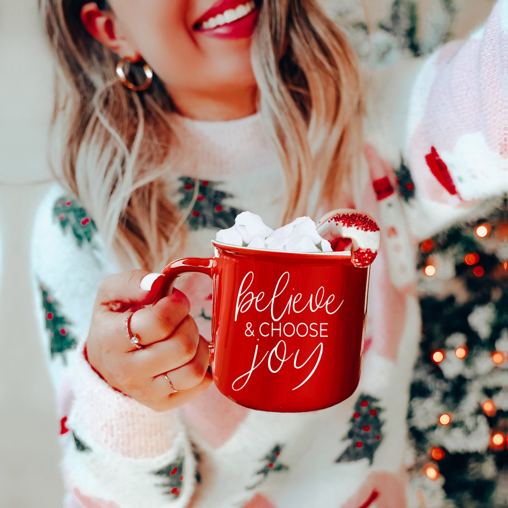 A ceramic Believe & Joy Mug in red with white lettering, featuring a positive message on both sides, perfect for holiday gifting.