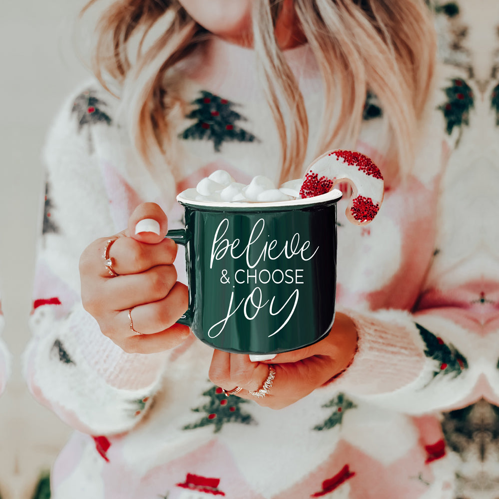 A ceramic Believe & Joy Mug in red with white lettering, featuring a positive message on both sides, perfect for holiday gifting.