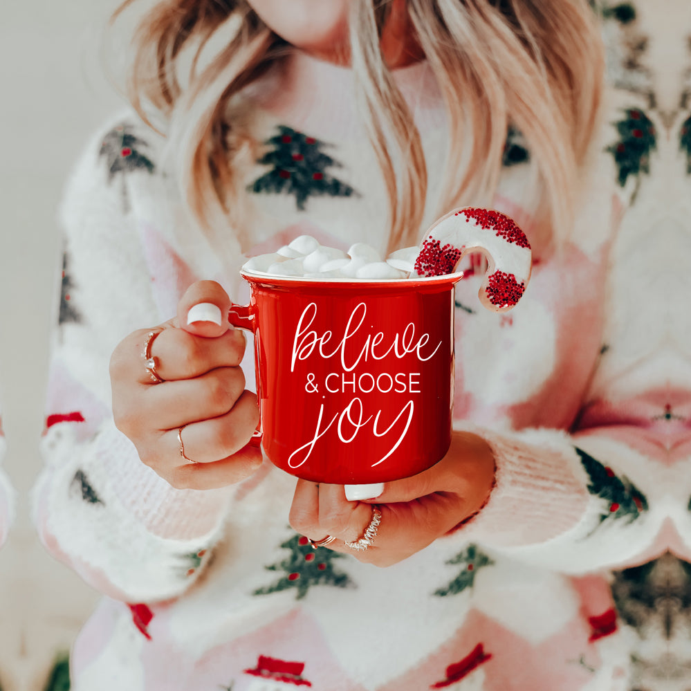 A ceramic Believe & Joy Mug in red with white lettering, featuring a positive message on both sides, perfect for holiday gifting.