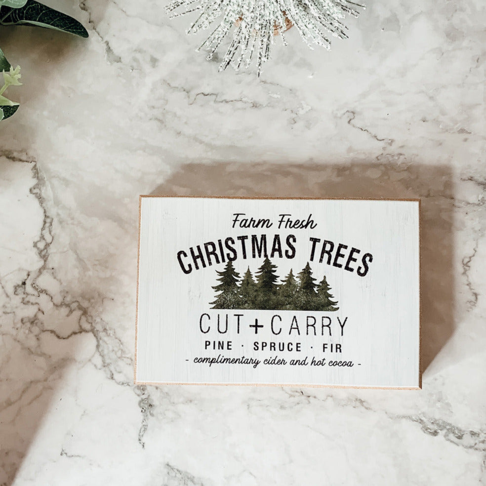 A decorative Cut + Carry Sign featuring a light gray textured background with the text about farm fresh Christmas trees and complimentary cider and hot cocoa.