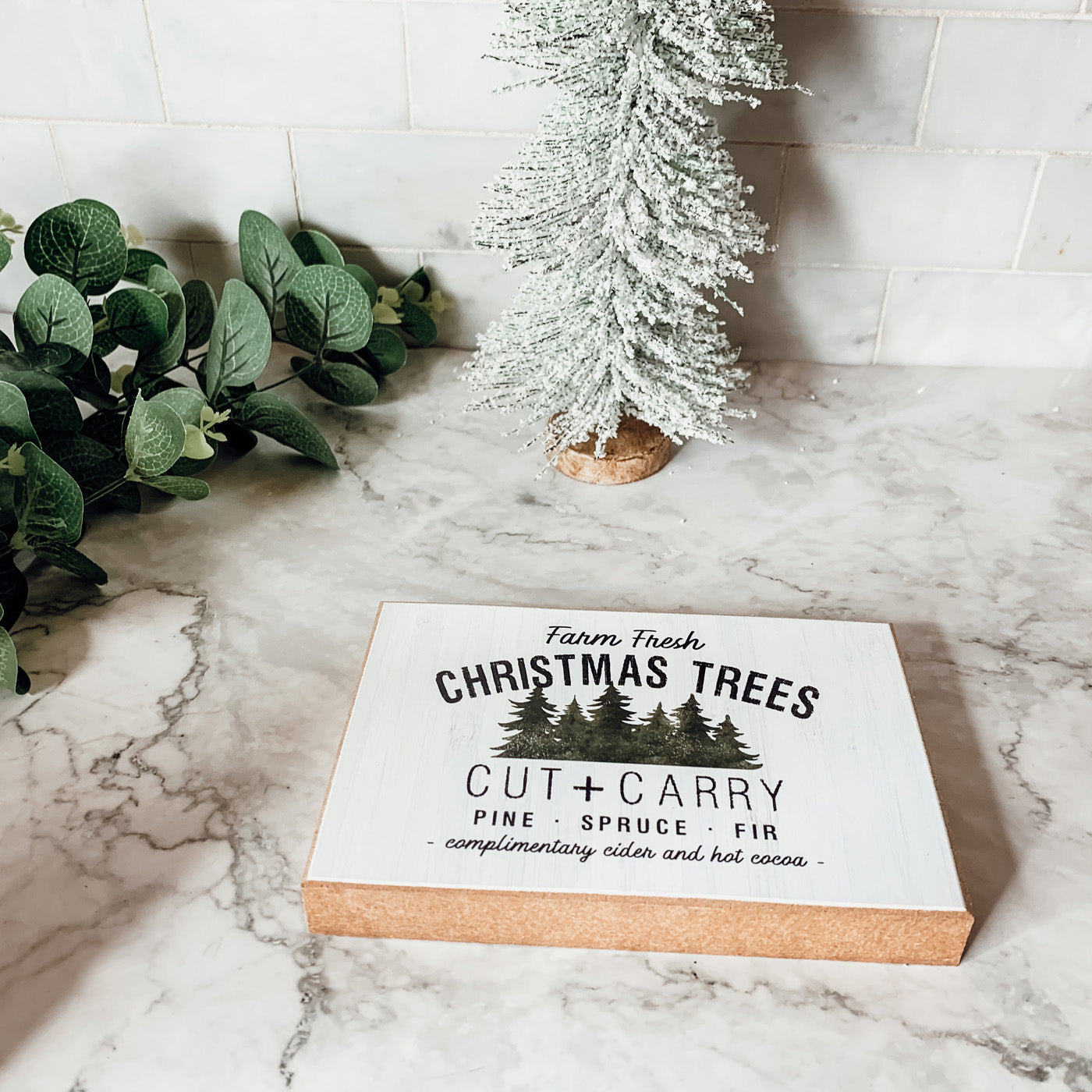 A decorative Cut + Carry Sign featuring a light gray textured background with the text about farm fresh Christmas trees and complimentary cider and hot cocoa.