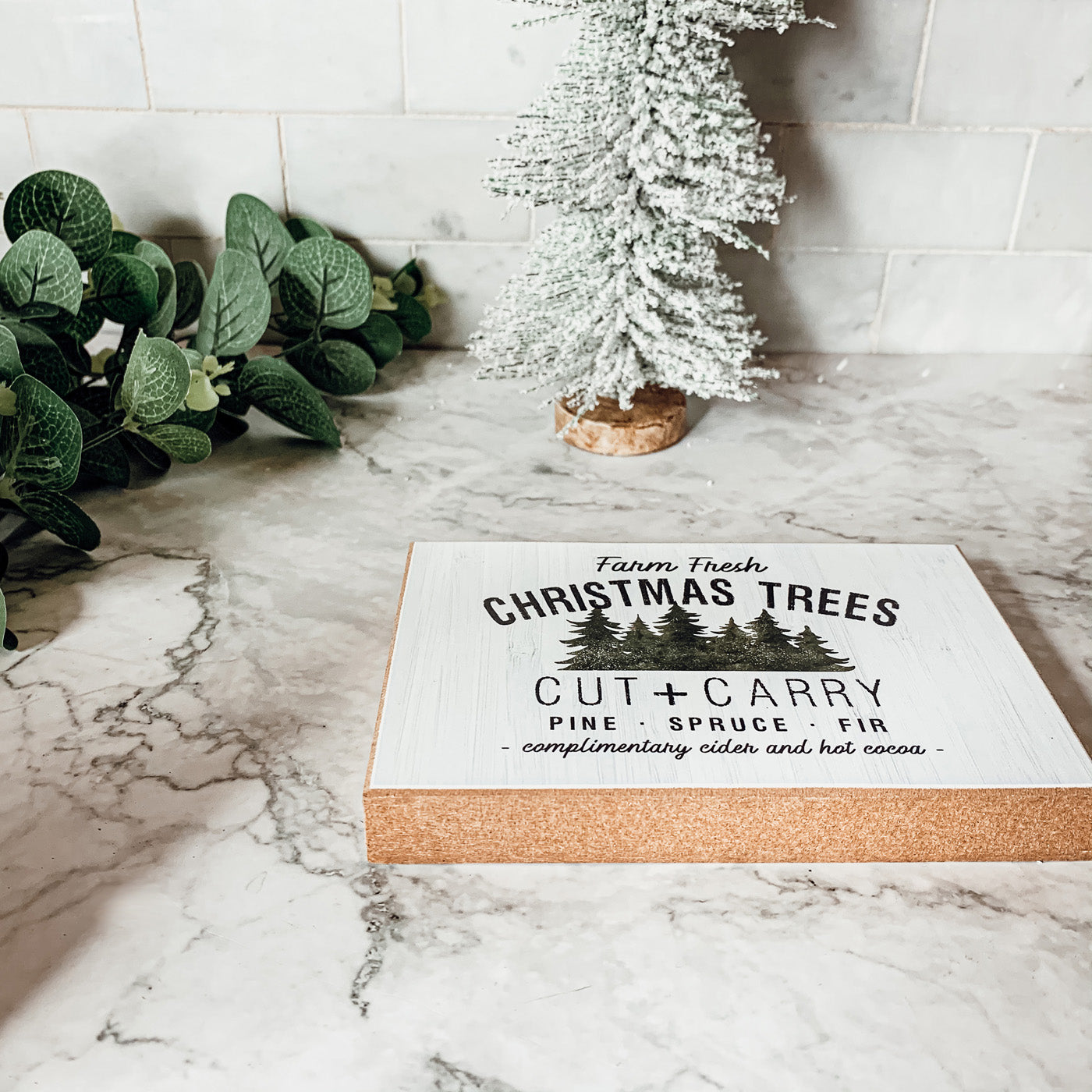 A decorative Cut + Carry Sign featuring a light gray textured background with the text about farm fresh Christmas trees and complimentary cider and hot cocoa.