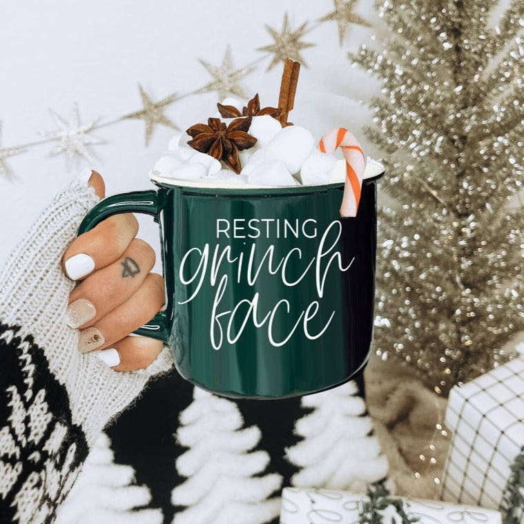 A whimsical Grinch Face Mug in red ceramic, featuring the Grinch's face and white lettering, perfect for holiday beverages.