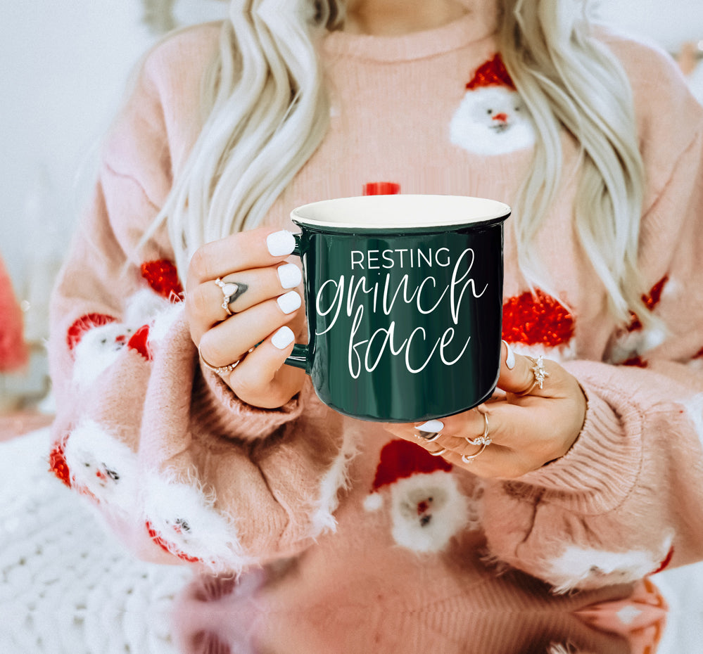 A whimsical Grinch Face Mug in red ceramic, featuring the Grinch's face and white lettering, perfect for holiday beverages.