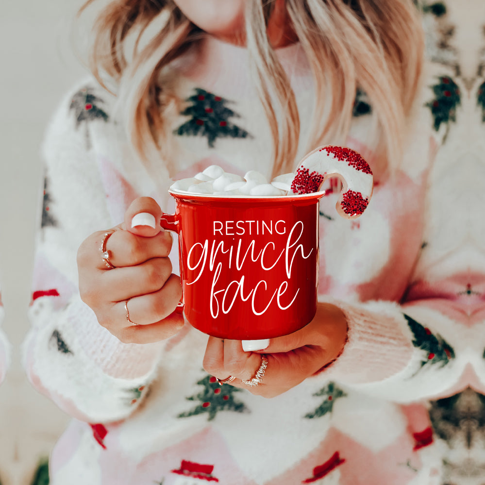 A whimsical Grinch Face Mug in red ceramic, featuring the Grinch's face and white lettering, perfect for holiday beverages.