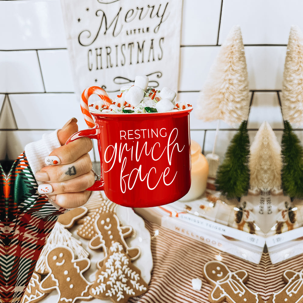 A whimsical Grinch Face Mug in red ceramic, featuring the Grinch's face and white lettering, perfect for holiday beverages.