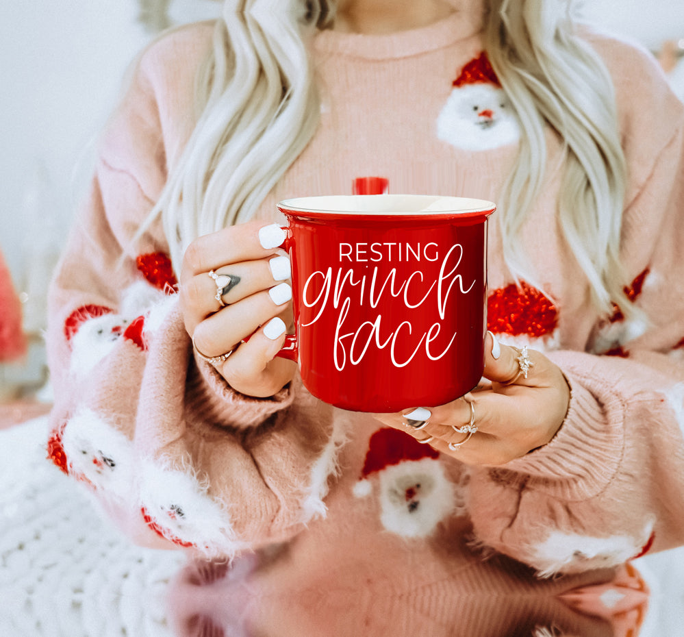 A whimsical Grinch Face Mug in red ceramic, featuring the Grinch's face and white lettering, perfect for holiday beverages.